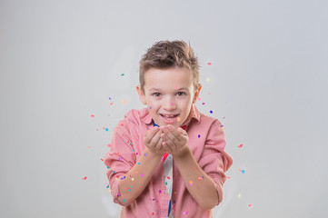 Сheerful brunette boy blows to confetti over grey background, concept of children's emotions and holiday