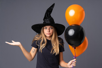 Young witch holding black and orange air balloons having doubts with confuse face expression