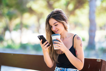 Young girl at outdoors holding coffee to take away while sending a message with the mobile