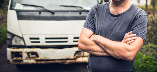 Male driver in old-fashioned t-shirt has crossed hands and stays near his old rusty truck on the...