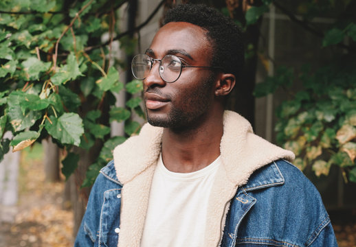 Portrait close-up modern african man thinking and looking away wearing eyeglasses in autumn city park on background of tree leaves