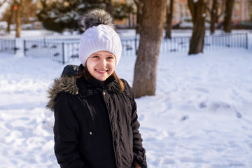 Beautiful young girl in a stylish warm white hat and in a dark coat walks in the city in winter where there is snow everywhere. Girl happy and smiling