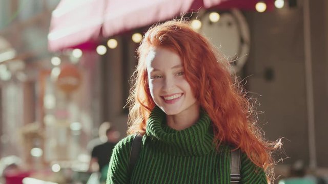 Outdoor portrait of nice-looking charming ginger girl with natural freckles and red hair posing in the street on lovely sunny day. Positive person, facial expression.