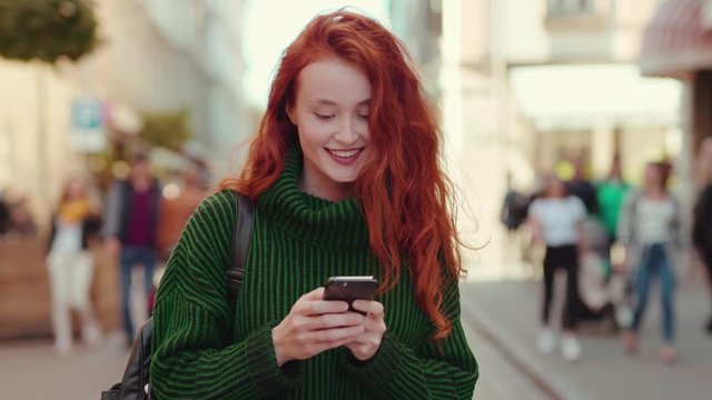 shot on Arra Alexa Mini Positive ginger girl student with charming smile walking in the street browsing smartphone content outdoor.