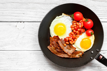 English breakfast in frying pan with fried eggs, bacon, beans and tomatoes on white wooden background