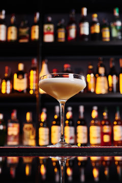Elegant female yellow cocktail with white foam, decorated with pink flower petal on dark table against the background of blurred bar with expensive alcohol. Close-up. Space for inscriptions