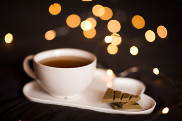 Evening cup of tea with chocolate bar on white plate closeup over glowing lights. Good morning. Breakfast.