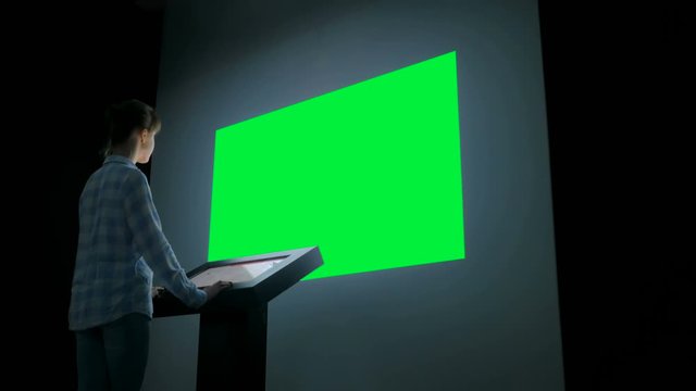 Green screen, mock up, futuristic, chroma key, template concept. Woman using electronic kiosk and looking at green blank large interactive wall display in dark room of modern technology museum