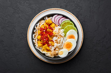 Nan Gyi Thoke in black bowl at dark slate background. Nan gyi Thohk is popular burmese cuisine dish with rice noodles, chicken breasts with spices, eggs, peanuts, lime and red onion. Top view