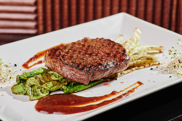 Juicy grilled steak on a pillow of Chinese cabbage grilled, served with red sauce and microgreen on a white square plate. Beautiful golden background. Close-up. Space.
