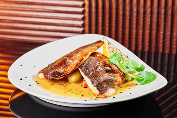 White steak of grilled fish on pillow of vegetables and yellow sauce, with lemon slice and fresh herbs. Fish on white plate on dark beautiful background. Close-up. Space