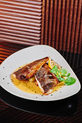 White steak of grilled fish on pillow of vegetables and yellow sauce, with lemon slice and fresh herbs. Fish on white plate on dark beautiful background. Close-up. Space