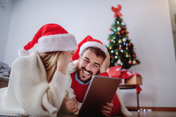 Cute Caucasian couple lying on stomach on the floor with santa hats on heads and using tablet for sending messages. Living room interior. In background is christmas tree.