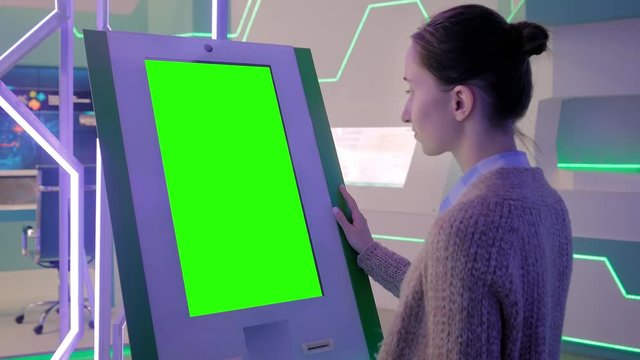 Woman looking at vertical digital interactive green display kiosk at exhibition or museum with futuristic sci-fi interior. Mock up, copyspace, template, chroma key, green screen, technology concept