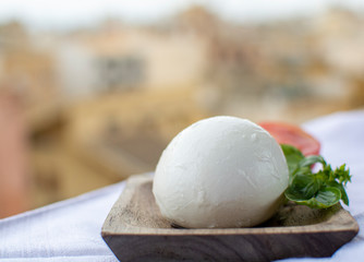 Italian mozzarella soft cheese ball served with basil and tomato on wooden plank with yellow houses in old Italian town on background