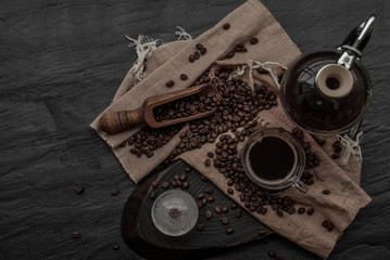 Cup of coffee and coffee beans roating with old wooden scoop and coffee beans around on the wooden and dark stone background. Top view with copy space for your text.