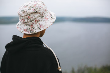 Hipster teenager in bucket hat standing on top of rock mountain and looking on river, view from back. Atmospheric calm moment. Wanderlust and travel. Copy space © sonyachny