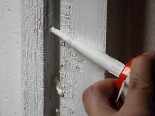 Application of white silicone sealant from a gun to an old painted vertical wooden surface