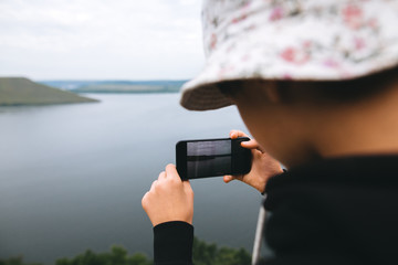 Hipster teenager streaming live while standing on top of rock mountain with amazing view on river. Young stylish guy exploring and traveling. Atmospheric moment. Copy space