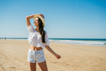 Gorgeous young girl with long dark hair and the straw hat on her head in a white sweater and shorts stands on the background of the sea coast and is surprised