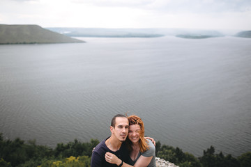 Happy hipster couple sitting on top of rock mountain with beautiful view on river. Tourist couple hugging and smiling on windy cliff. Traveling together. Copy space