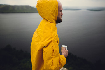 Traveler in yellow raincoat holding metal mug and standing on cliff  in rainy windy day with view...