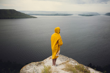 Fototapeta na wymiar Hipster traveler in yellow raincoat standing on cliff and looking at lake in windy moody day. Wanderlust and travel concept. Man hiking in Norway on foggy day. Atmospheric moment