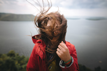 Traveler girl in red raincoat and with windy hair standing on top of rock mountain, enjoying beautiful view on river. Young hipster woman in glasses relaxing on cliff. Copy space. Explore - obrazy, fototapety, plakaty