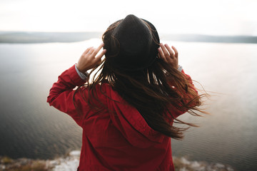 Traveler girl with windy hair holding hat and looking at evening sky on top of rock mountain,...