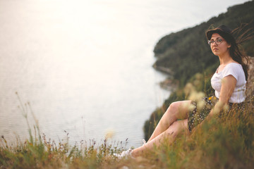 Traveler hipster girl in hat and windy hair sitting on top of rock mountain in grass, enjoying beautiful sunset view on river. Atmospheric moment. Copy space