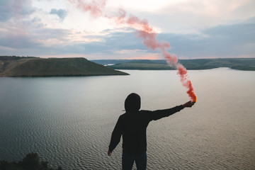 Obraz na płótnie Canvas Stylish hipster holding red smoke bomb in hand, standing on top of rock mountain with amazing sunset view on river. Atmospheric moment. Traveler guy with smoke. Copy space