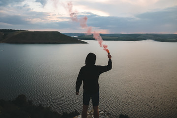 Ultras hooligan holding red smoke bomb in hand, standing on top of rock mountain with amazing...
