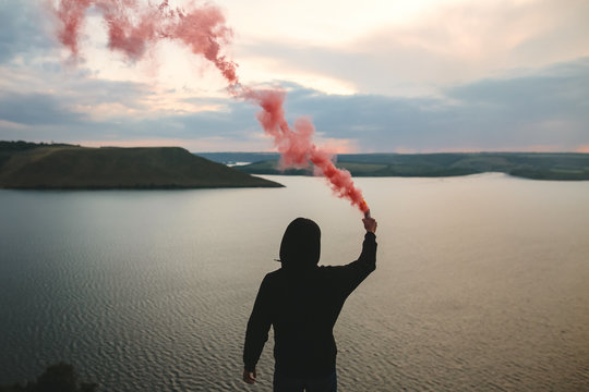 Ultras hooligan holding red smoke bomb in hand, standing on top of rock mountain with amazing sunset view on river. Atmospheric moment. Traveler guy with smoke. Copy space