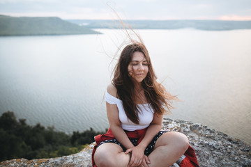 Hipster girl with windy hair sitting and meditating on top of rock mountain with beautiful view on...