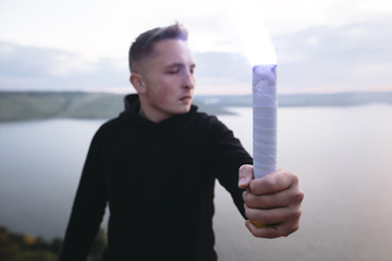 Ultras hooligan holding blue flare torch in hand, standing on top of rock mountain in evening with...