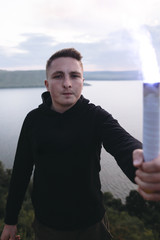 Fototapeta premium Protest concept. Brutal man holding blue flare torch in hand, standing on top of hill in evening with view on river. Ultras hooligan with fire signal bomb. Atmospheric moment