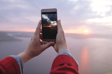 Traveler girl holding phone and taking photo of beautiful evening sky on top of rock mountain. Copy space. Atmospheric moment. Hipster girl making photo of sunset