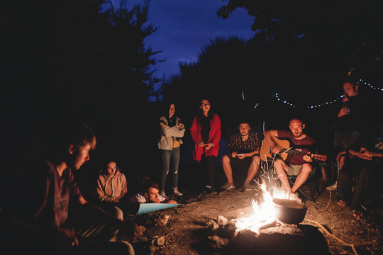 Hipster man playing on acoustic guitar and singing song with friends travelers at big bonfire at night camp in the forest. Group of people chilling at fire in the evening, camping near lake