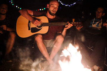 Obraz premium Friends travelers chilling at big bonfire, singing songs and playing guitar at camp in the night forest. Group of people resting at fire in the evening, camping in woods