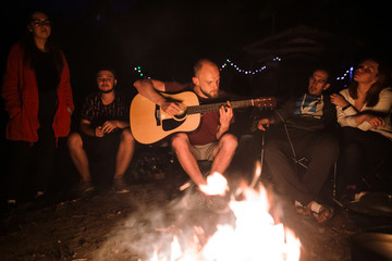 Friends travelers chilling at big bonfire, singing songs and playing guitar at camp in the night...