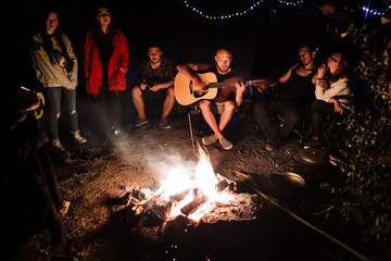 Friends travelers chilling at big bonfire, singing songs and playing guitar at camp in the night forest. Group of people resting at fire in the evening, camping in woods