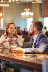 Cheerful long haired brunette woman sits a bar counter, holds a cup of tea, looks with tender smile at impressive man in bright lighted restaurant, close up, bottom shot