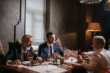 Charming young woman sits opposite to pleasant good looking men, talks kindly, feels positive emotions, people eat delicious meal at well served table in dark restaurant,