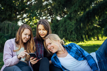 Three young girls sitting on green grass with book and smartphones. Cheerful friends resting in park. Spring and summer leisure on a nature, concept of female friendship.