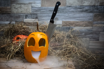 Yellow and funny halloween pumpkin with a knife in his head and smoke or steam from his mouth. Stands on a wooden stand against a stone wall. Red jack o lantern on background