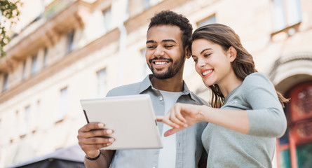 Beautiful happy couple using digital tablet. Joyful smiling woman and man looking at gadget in a city in summer. Love, relationship, technology and communication concept