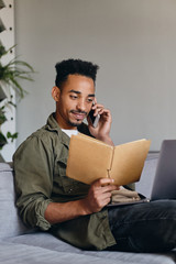 Young attractive smiling African American man with notebook happily talking on cellphone in co-working space