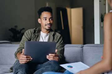 Young attractive casual African American man with laptop dreamily working with colleague in co-working space