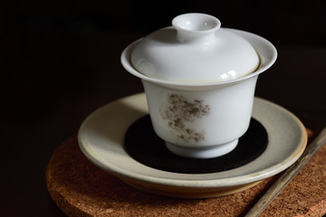 Close-up of an isolated Chinese white teapot with tea  and lid , which stands on a plate and on a cork coaster with a used chopsticks next to it against dark background with place at a tea ceremony