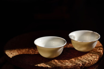 Fototapeta na wymiar Close up of two empty small white chinese tea cups standing against a dark background on a cork plate with free space for text at a Chinese tea ceremony